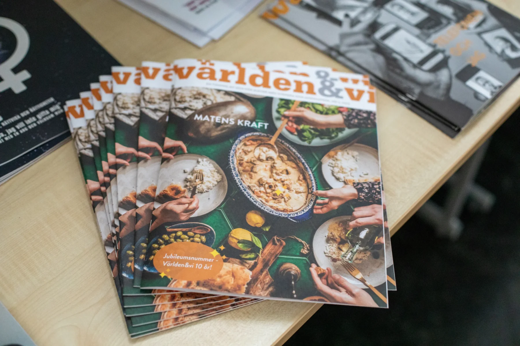 En bunt nummer av tidningen Världen & vi ligger på ett bord. Omslaget visar personer som äter tillsammans runt ett bord. Text på framsidan lyder Matens kraft och Jubiläumsnummer Världen&vi 10 år.