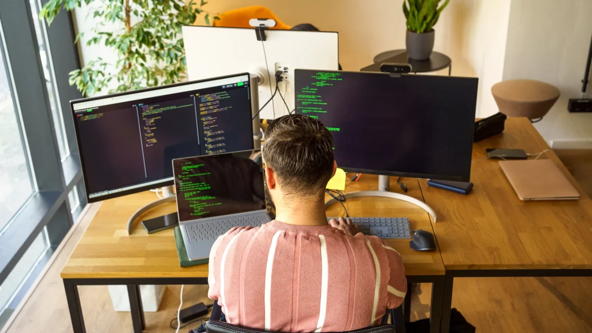 En person sitter vid ett skrivbord och arbetar med kod på tre skärmar samt en bärbar dator. Arbetsplatsen är ljus med stora fönster, växter och ordnade skrivbord runtomkring.