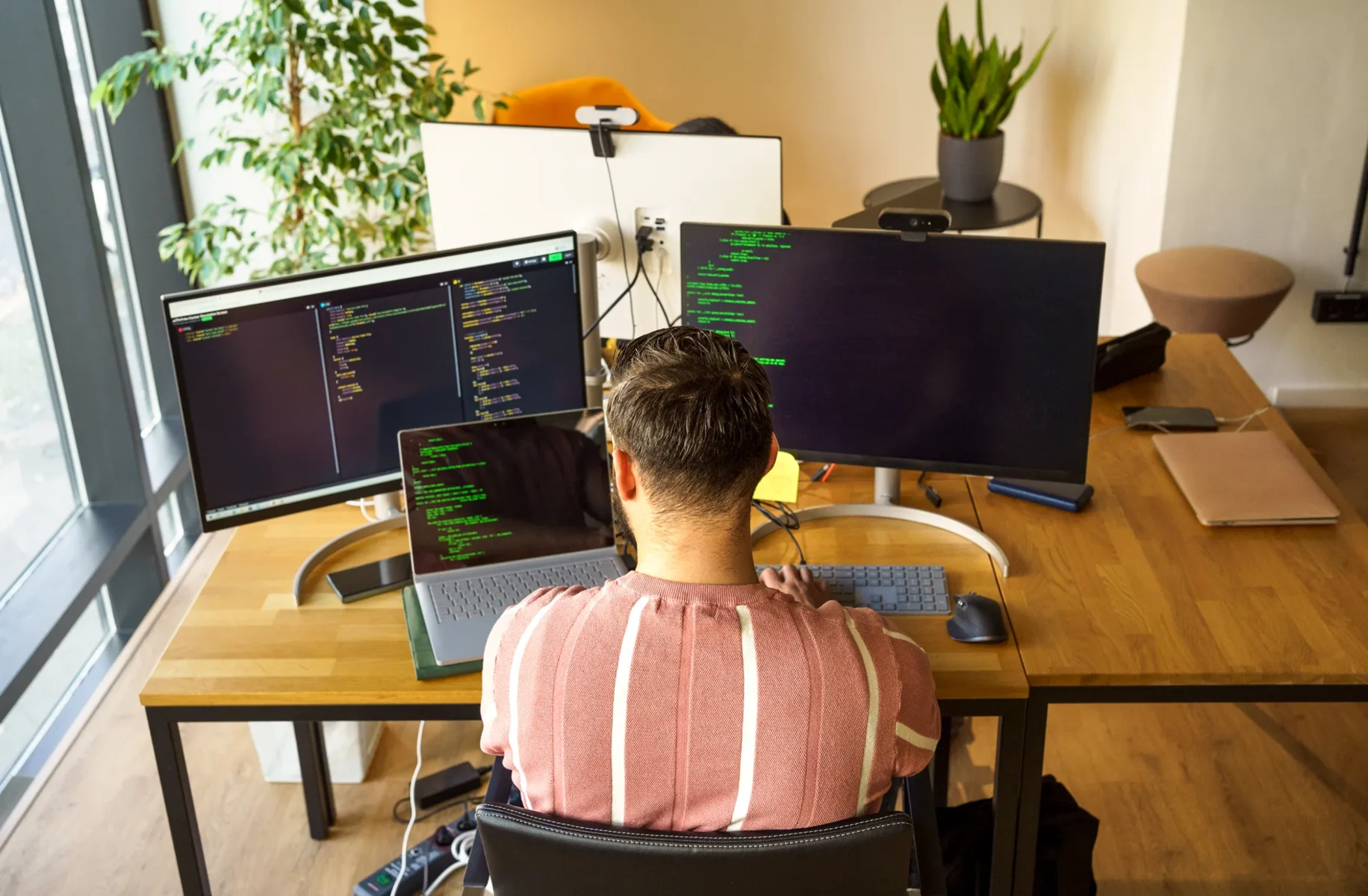 En person sitter vid ett skrivbord och arbetar med kod på tre skärmar samt en bärbar dator. Arbetsplatsen är ljus med stora fönster, växter och ordnade skrivbord runtomkring.