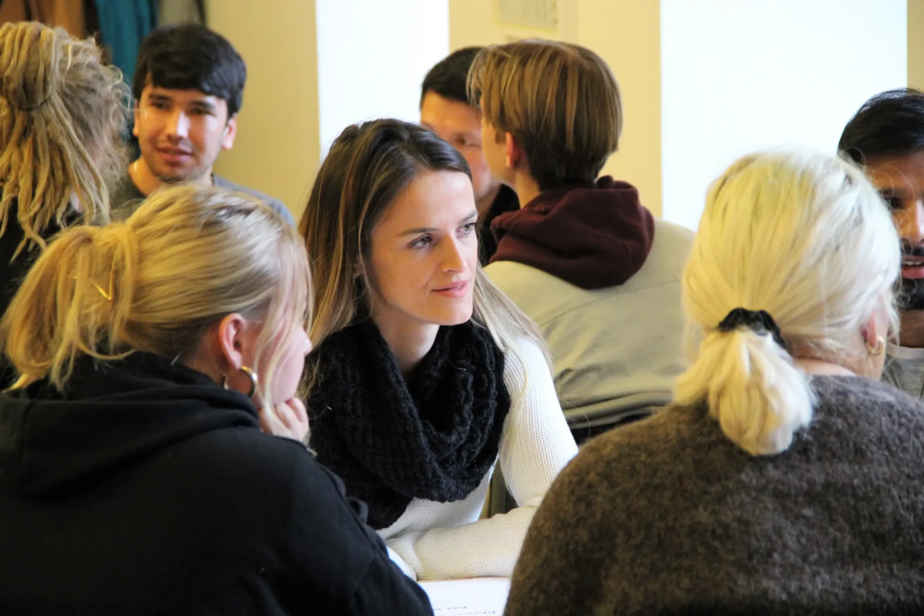 En grupp människor sitter i ett rum och samtalar i mindre grupper. I förgrunden syns en kvinna i vit tröja och svart halsduk som lyssnar på dem hon sitter med. I bakgrunden syns fler deltagare som pratar med varandra.