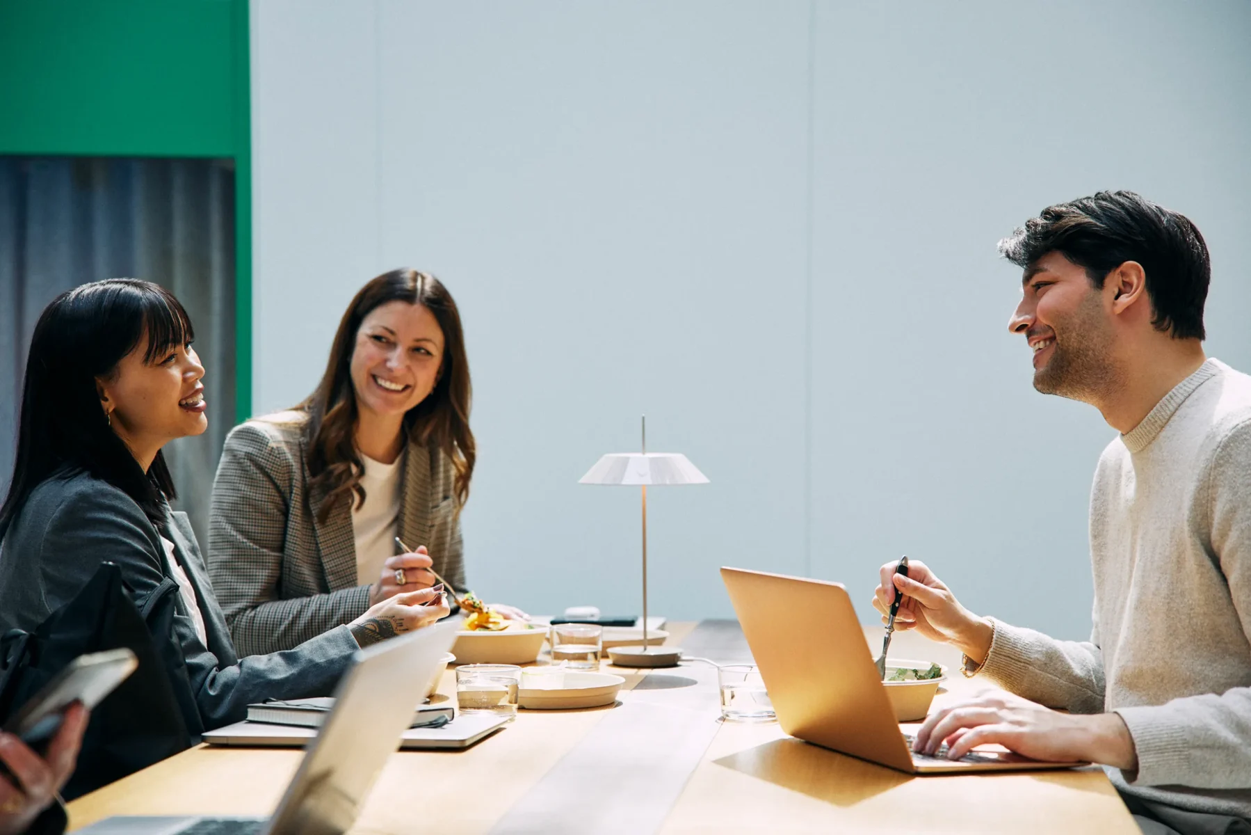 Tre personer sitter vid ett långt bord i en ljus miljö och äter samtidigt som de arbetar vid sina laptops. De samtalar och ler mot varandra. På bordet står en liten lampa, glas och skålar med mat.