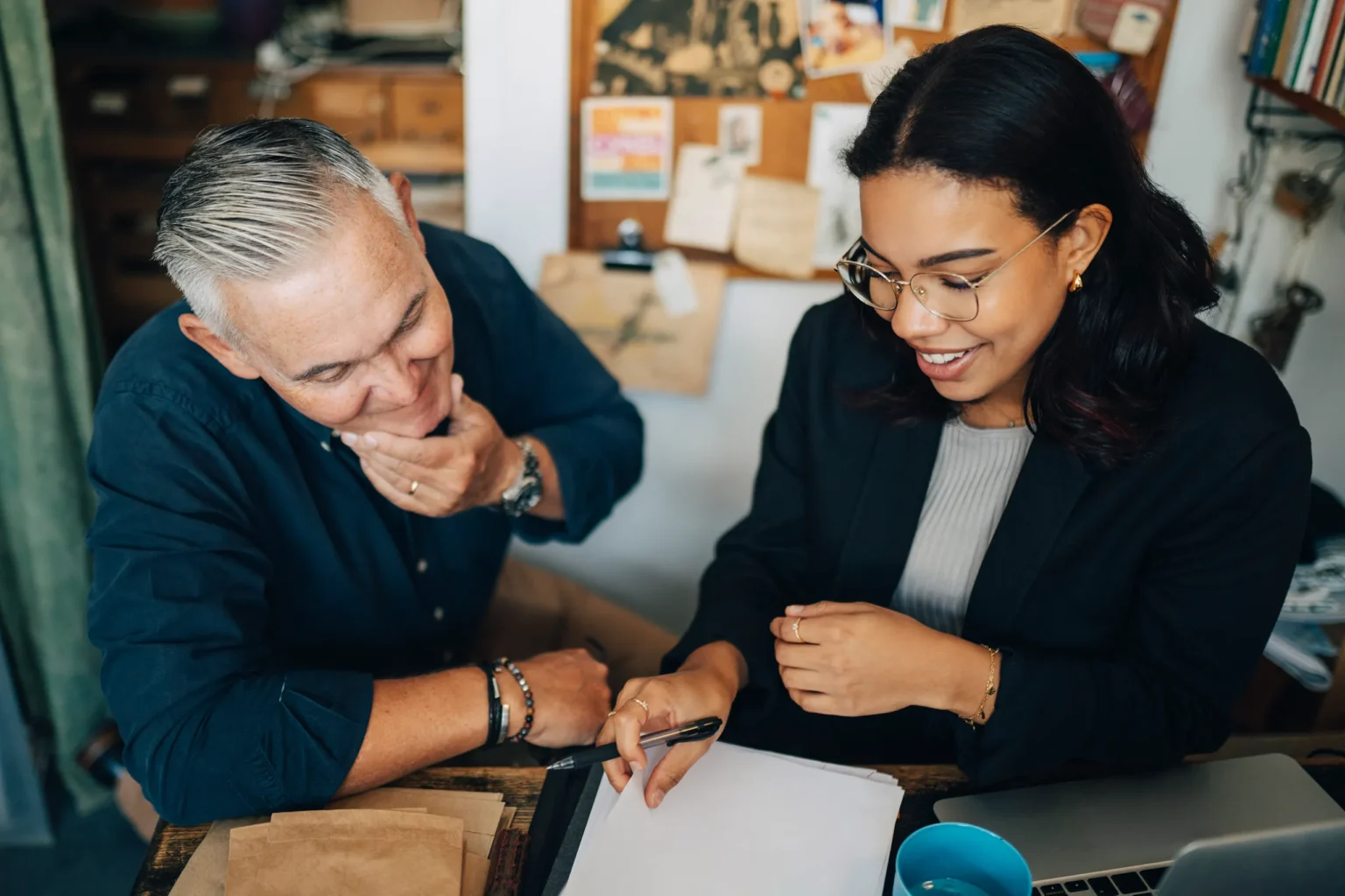 En äldre man och en yngre kvinna sitter vid ett bord och går igenom dokument tillsammans. Kvinnan håller en penna och ler medan mannen lyssnar och tittar ner på papperen. På bordet ligger en laptop och kuvert.