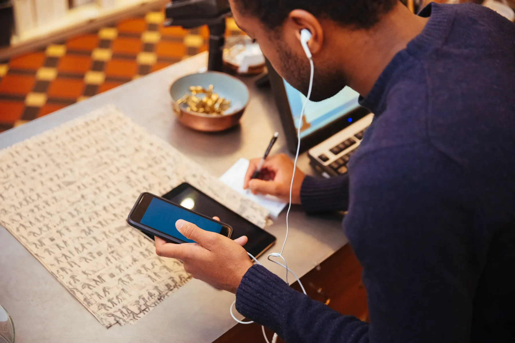 En person sitter vid ett bord och arbetar med både mobiltelefon och laptop samtidigt. Hen skriver anteckningar på ett papper medan hörlurar är inkopplade. På bordet finns även en skål med små föremål och en surfplatta.