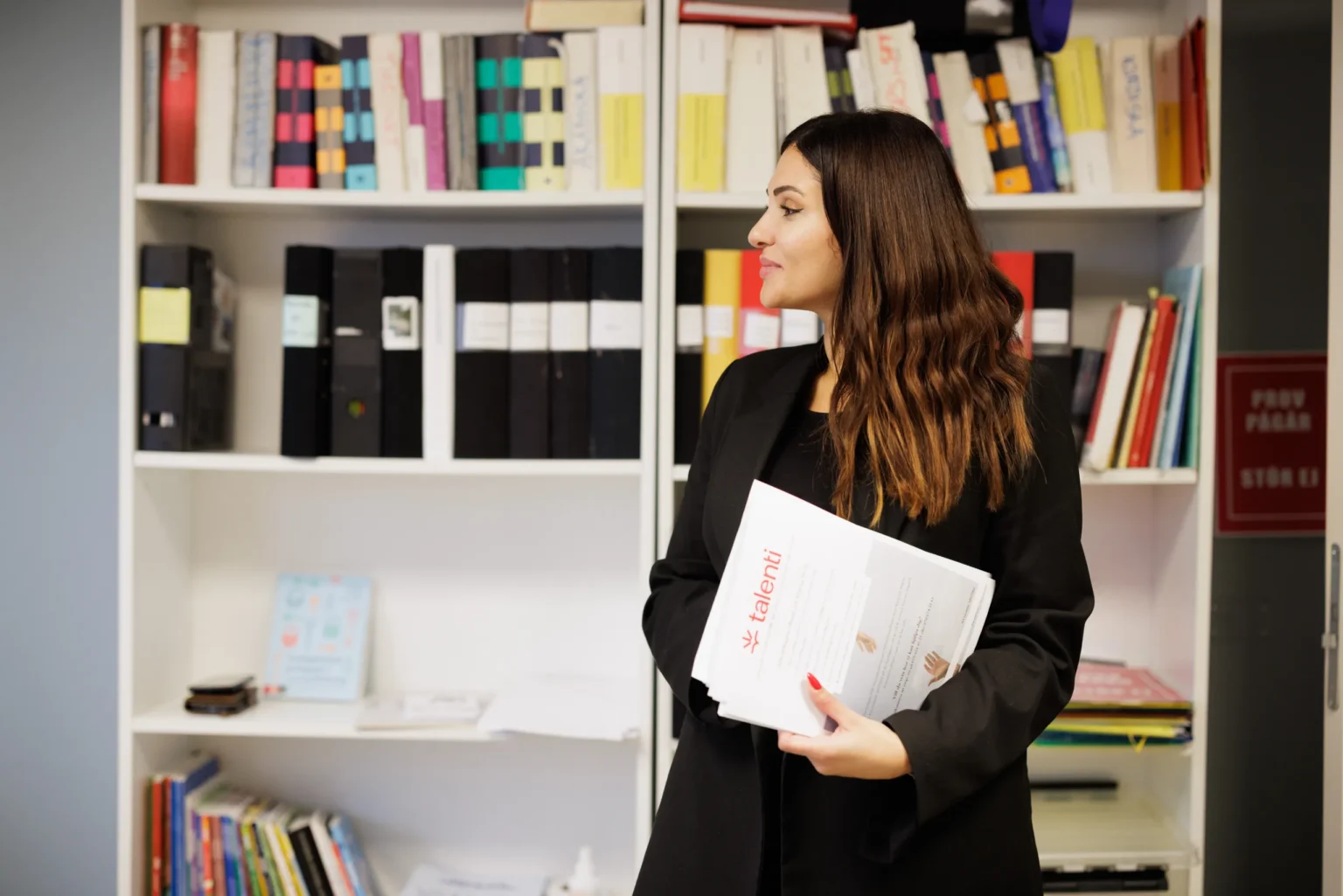 En kvinna står framför en bokhylla fylld med pärmar och böcker. Hon håller dokument med logotypen “Talenti” i famnen och tittar åt sidan med ett lugnt uttryck.