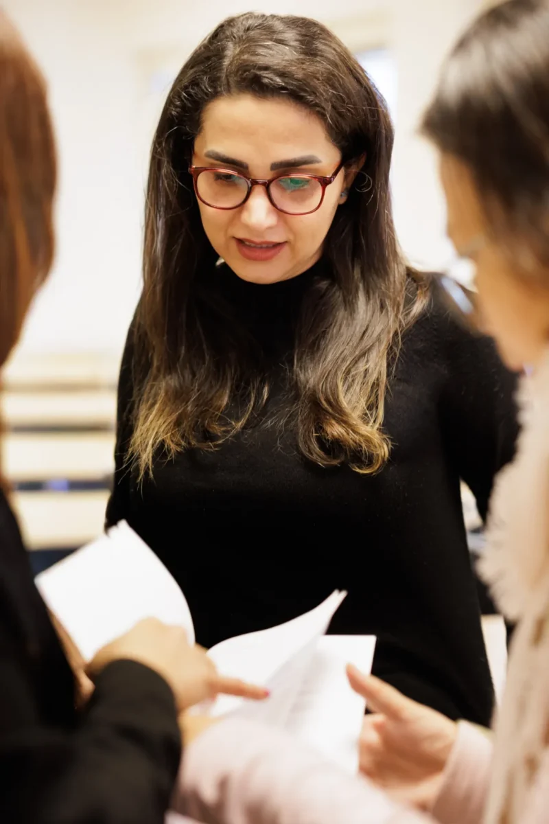 En kvinna med långt mörkt hår och glasögon står i ett rum och tittar ned på papper som två andra personer håller framför henne.