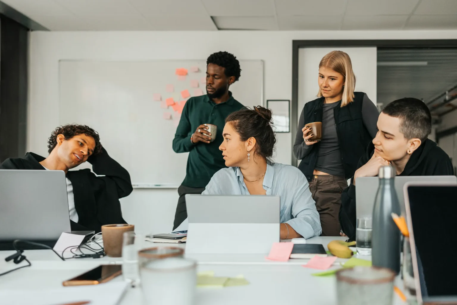 En grupp unga vuxna arbetar tillsammans runt ett bord med laptops, anteckningar och kaffekoppar, medan några står och håller muggar och pratar framför en whiteboard med post it-lappar.