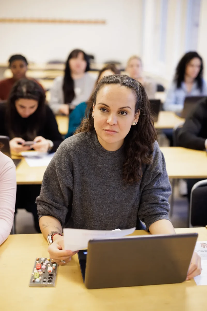 En kvinna sitter i ett klassrum framför en laptop och håller i papper, medan andra deltagare arbetar i bakgrunden.