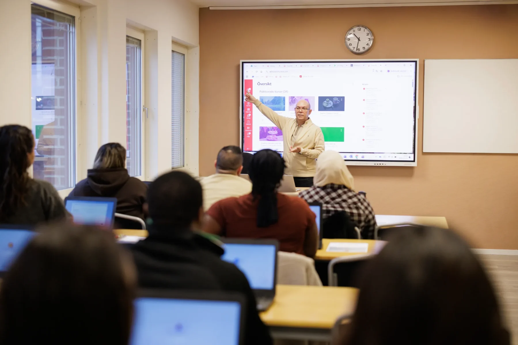 En lärare står framför en skärm och pekar på en presentation i ett klassrum där deltagare sitter med laptops och följer undervisningen.
