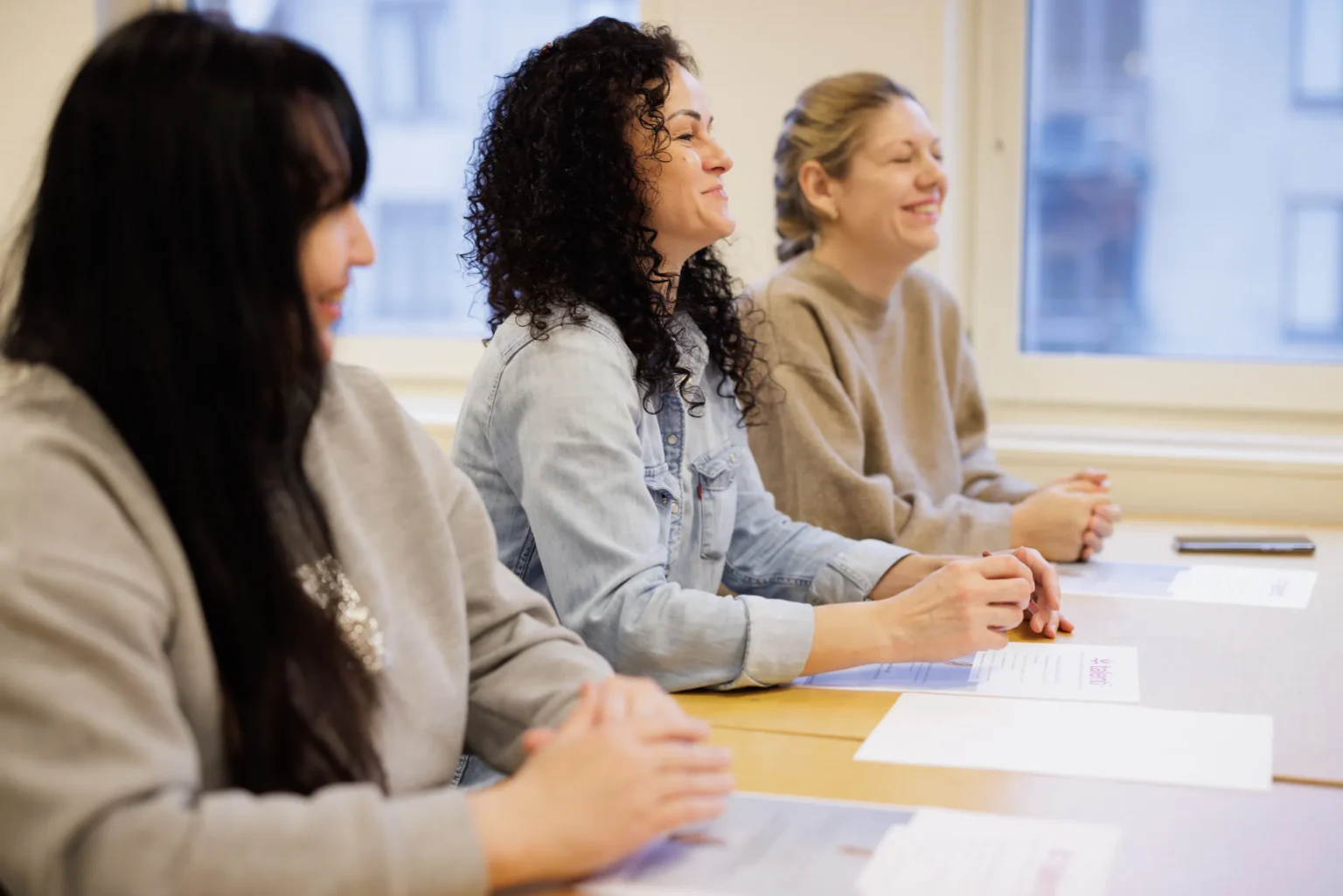 Tre kvinnor sitter vid ett bord i ett klassrum och lyssnar leende, med papper framför sig och ett fönster i bakgrunden.