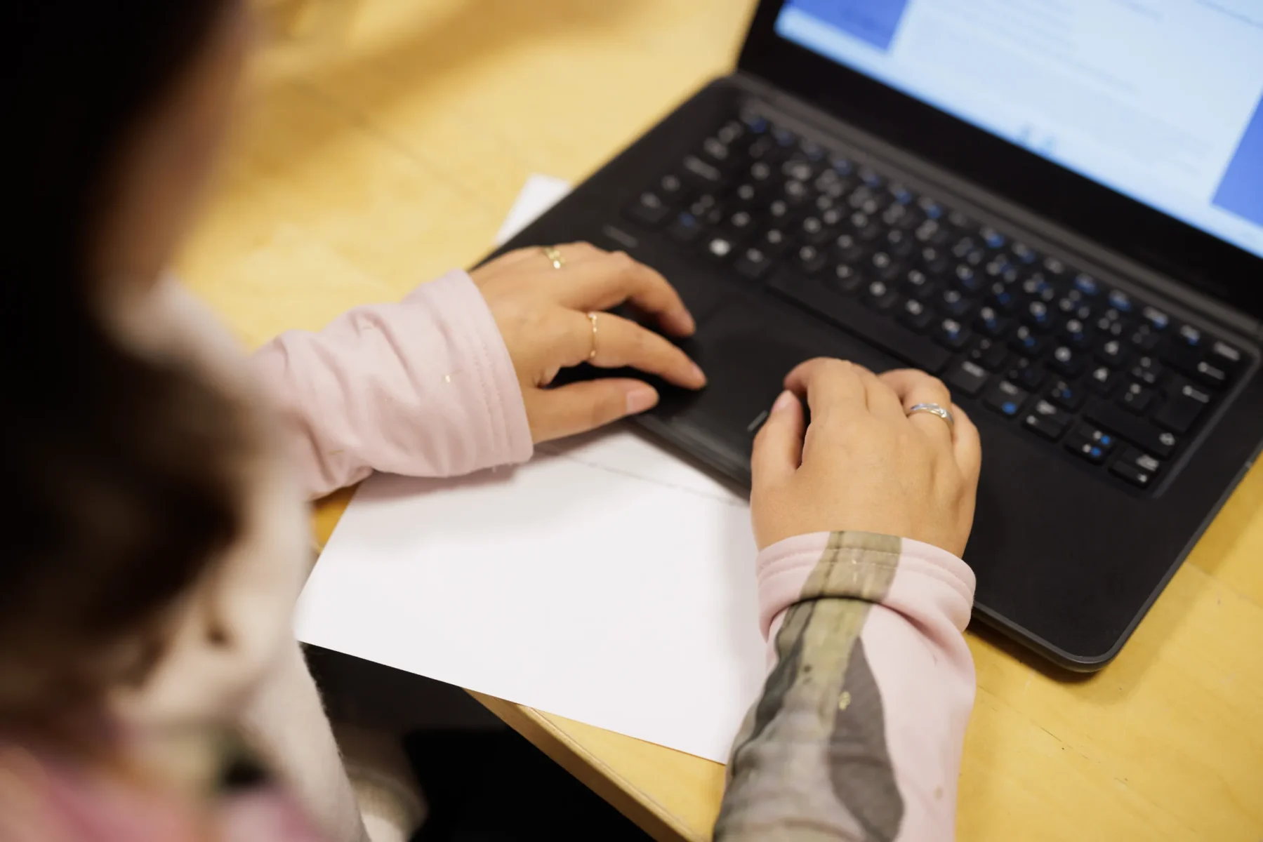En person sitter vid ett bord och skriver på en bärbar dator, med händerna placerade över tangentbordet och ett papper framför sig.