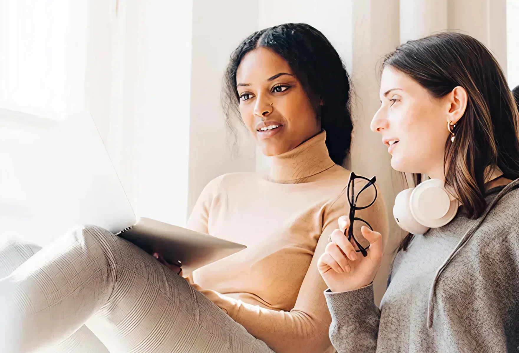 Två personer sitter bredvid varandra vid ett ljust fönster och tittar på en bärbar dator. Den ena håller glasögon i handen medan de samtalar och fokuserar på skärmen.